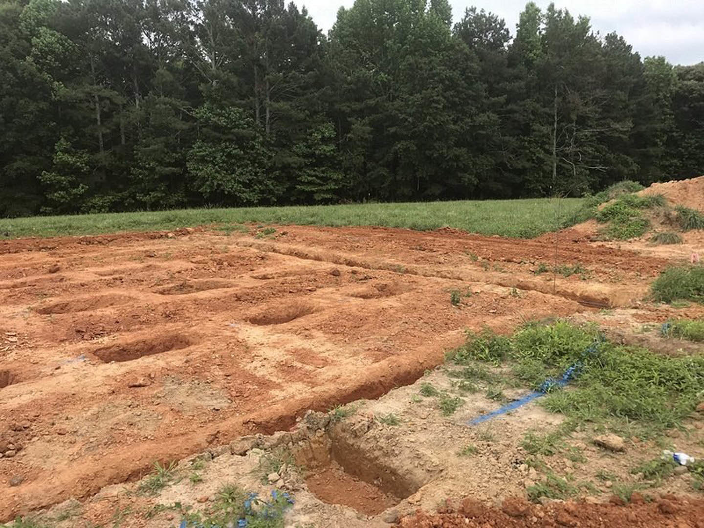 Dirt field with square holes, blue line marking in grass, group of trees in background under open sky