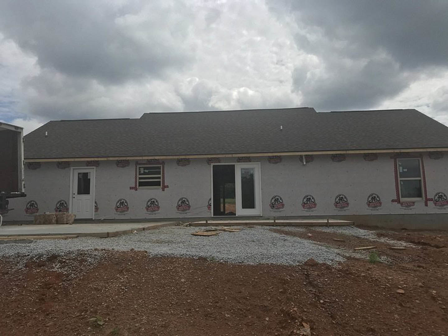Partially built house with exposed framing, white door with glass window, gravel and dirt foreground, overcast sky with dense clouds