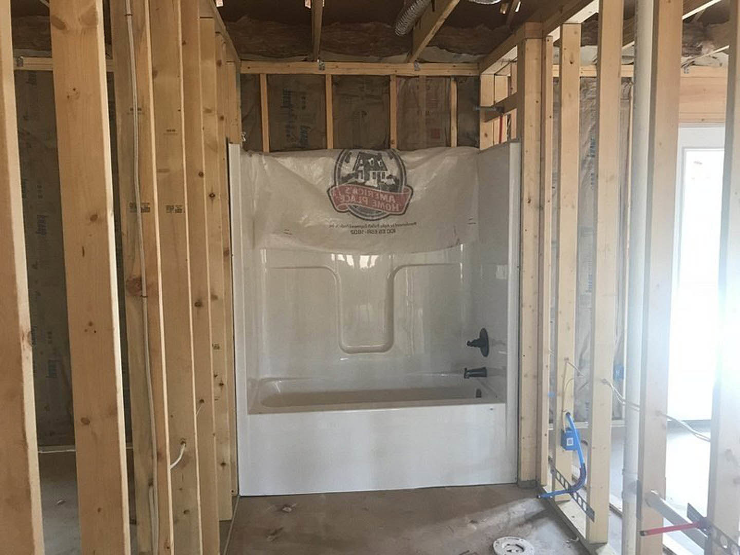 Bathroom under construction with exposed wood framing, plastic covering over shower area, white bathtub, visible building insulation, and unfinished walls and ceiling.