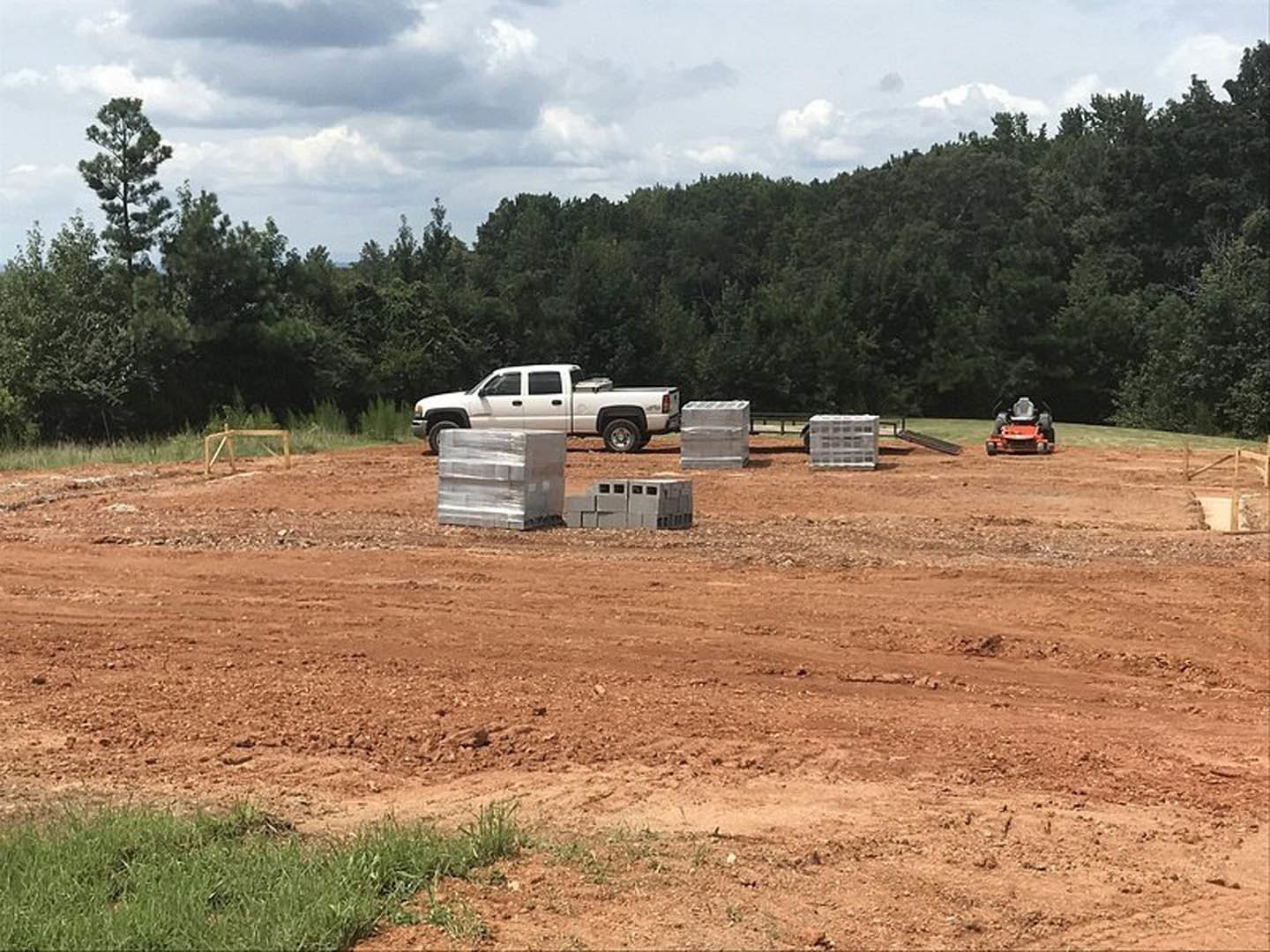 White pickup truck parked on a dirt lot beside a pile of cardboard boxes, person riding a lawnmower in the background, grassy field bordered by trees under partly cloudy sky.