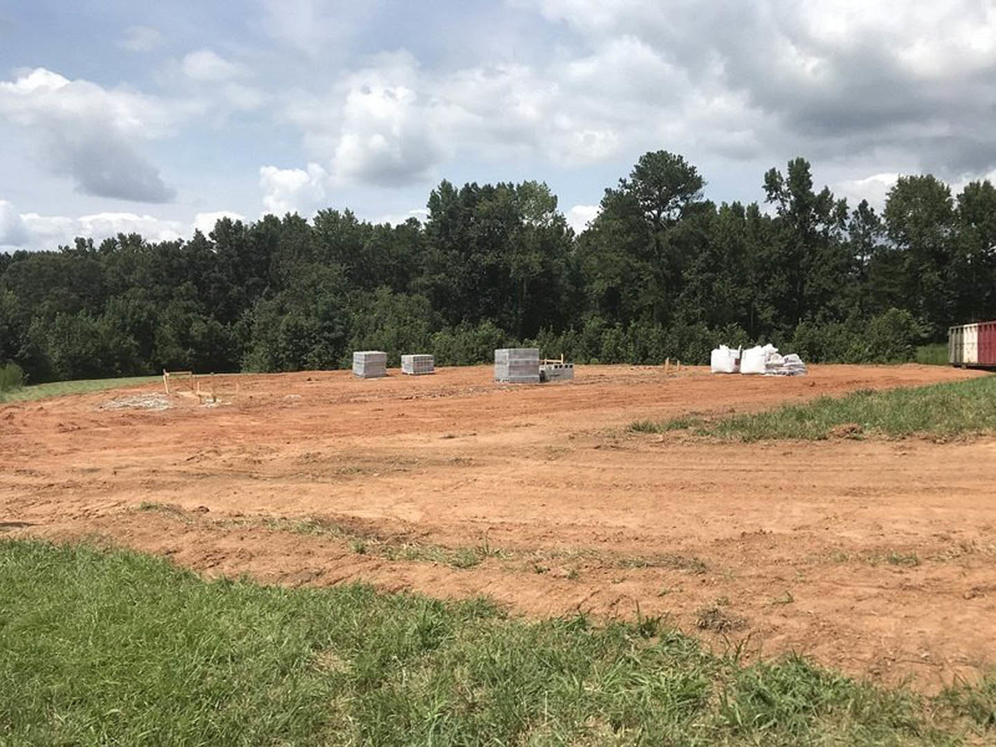 Dirt field with white construction blocks, scattered bricks, and trees under a partly cloudy blue sky