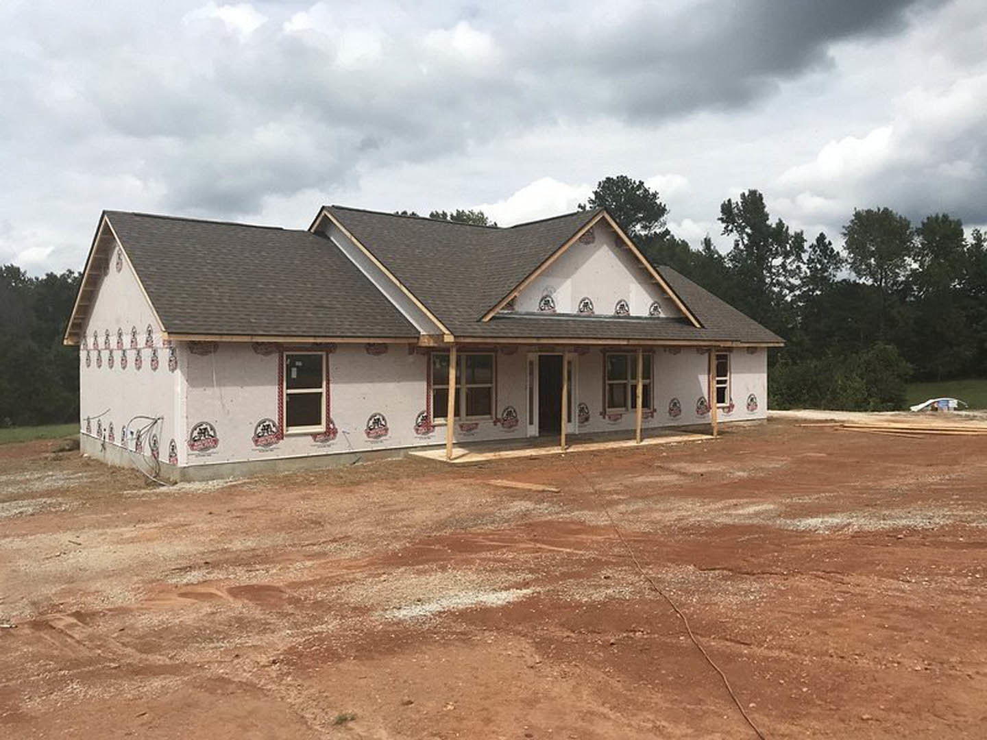Two-story house under construction with exposed dirt yard, red front door, white-framed windows, glass panes, and trees in the background under a cloudy sky
