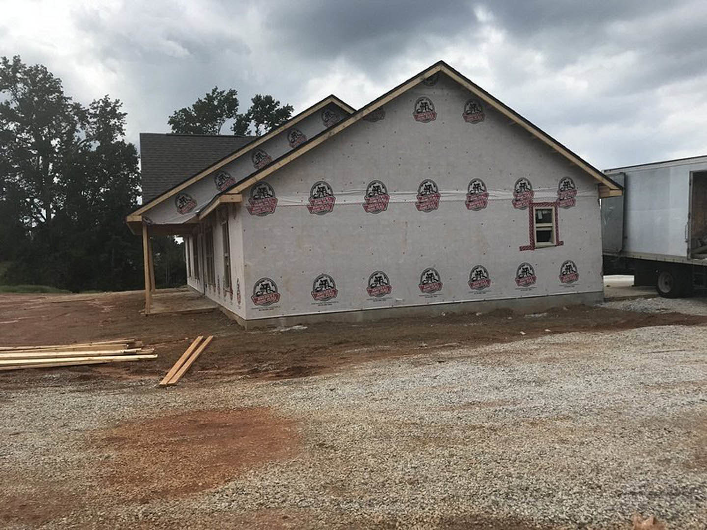 Partially built house with taped exterior walls, shingled roof, open white truck parked nearby, dirt construction site surrounded by leafy trees, black and white logo visible on