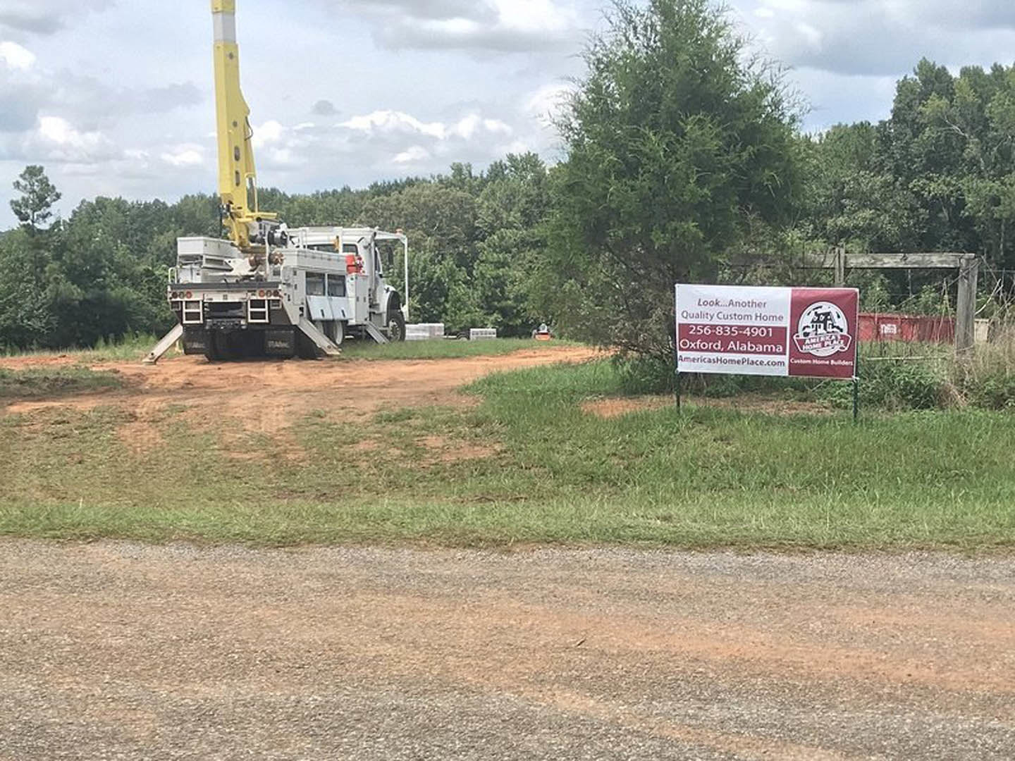 White pickup truck parked on dirt driveway in front of grassy land lot, billboard sign and yellow tower with flag in background, scattered trees under partly cloudy sky.