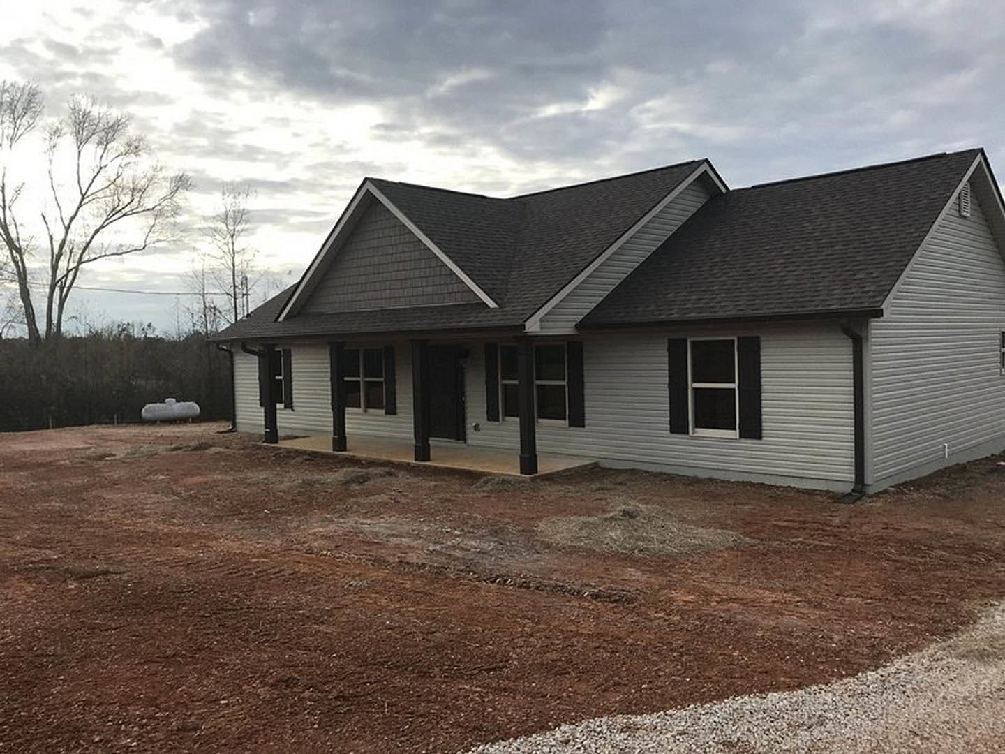 Modern home under construction featuring a black roof, white-framed windows, exposed dirt foundation, and leafless trees surrounding the property