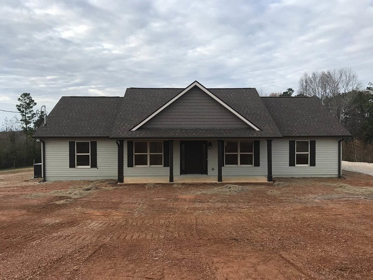 Two-story house with white siding, black front door framed in white, dirt driveway, cloudy sky overhead, and trees in the background.