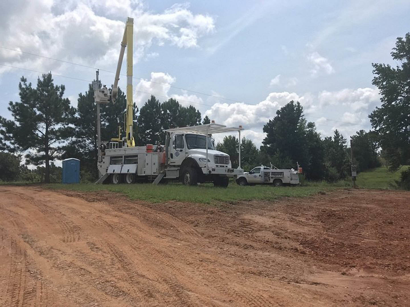 White truck with open door and yellow flag parked on dirt road beside blue portable toilet; cars and tall yellow and white pole in background, surrounded by trees and cloudy sky.