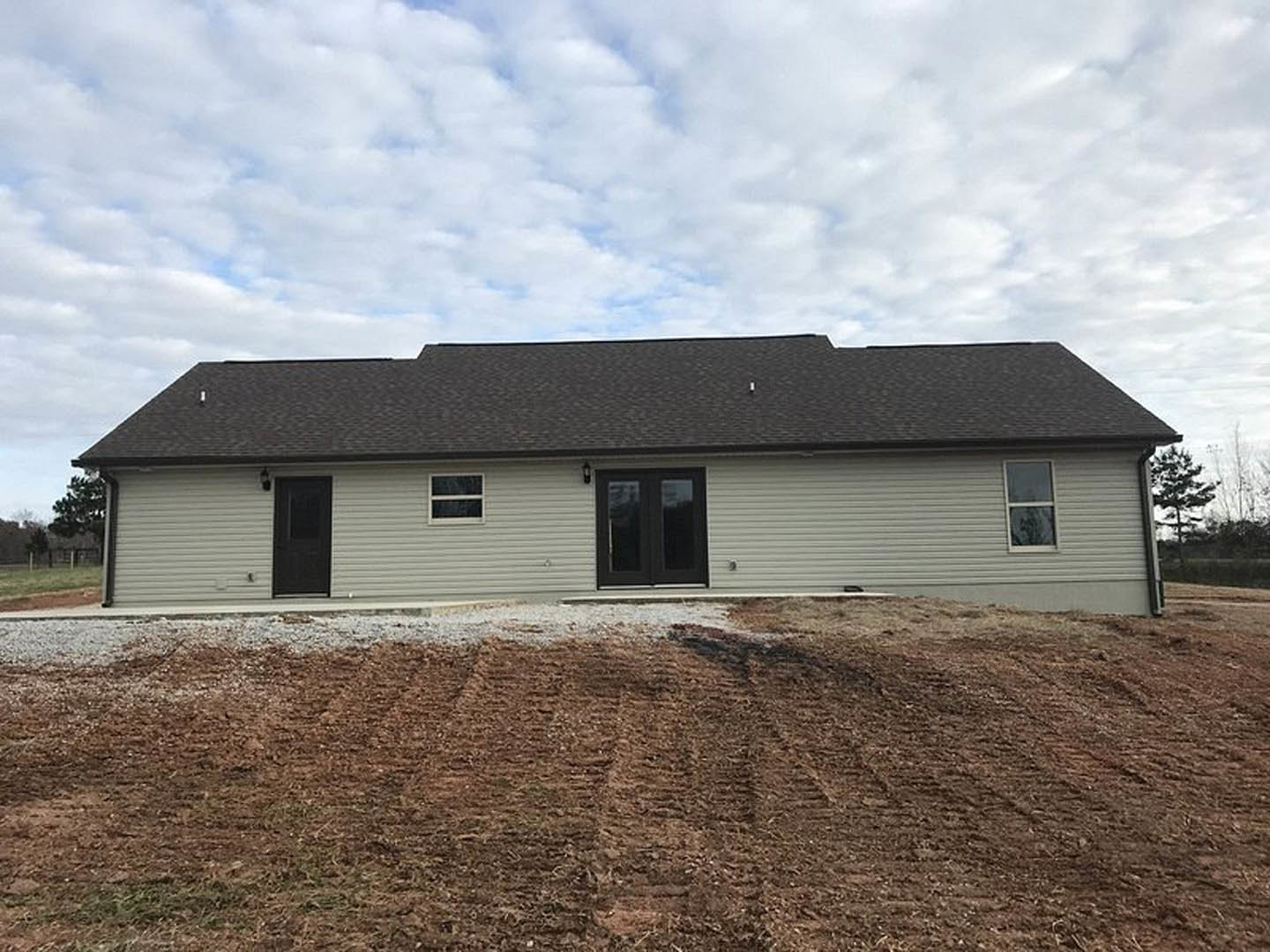 Modern house with light siding, black front door featuring a window, large window on facade, dirt field with tire tracks in foreground, cloudy sky above