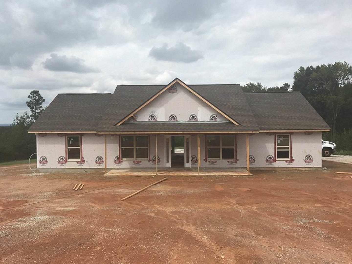 Partially built house with exposed framing and roof, surrounded by dirt road and construction materials, cloudy sky overhead