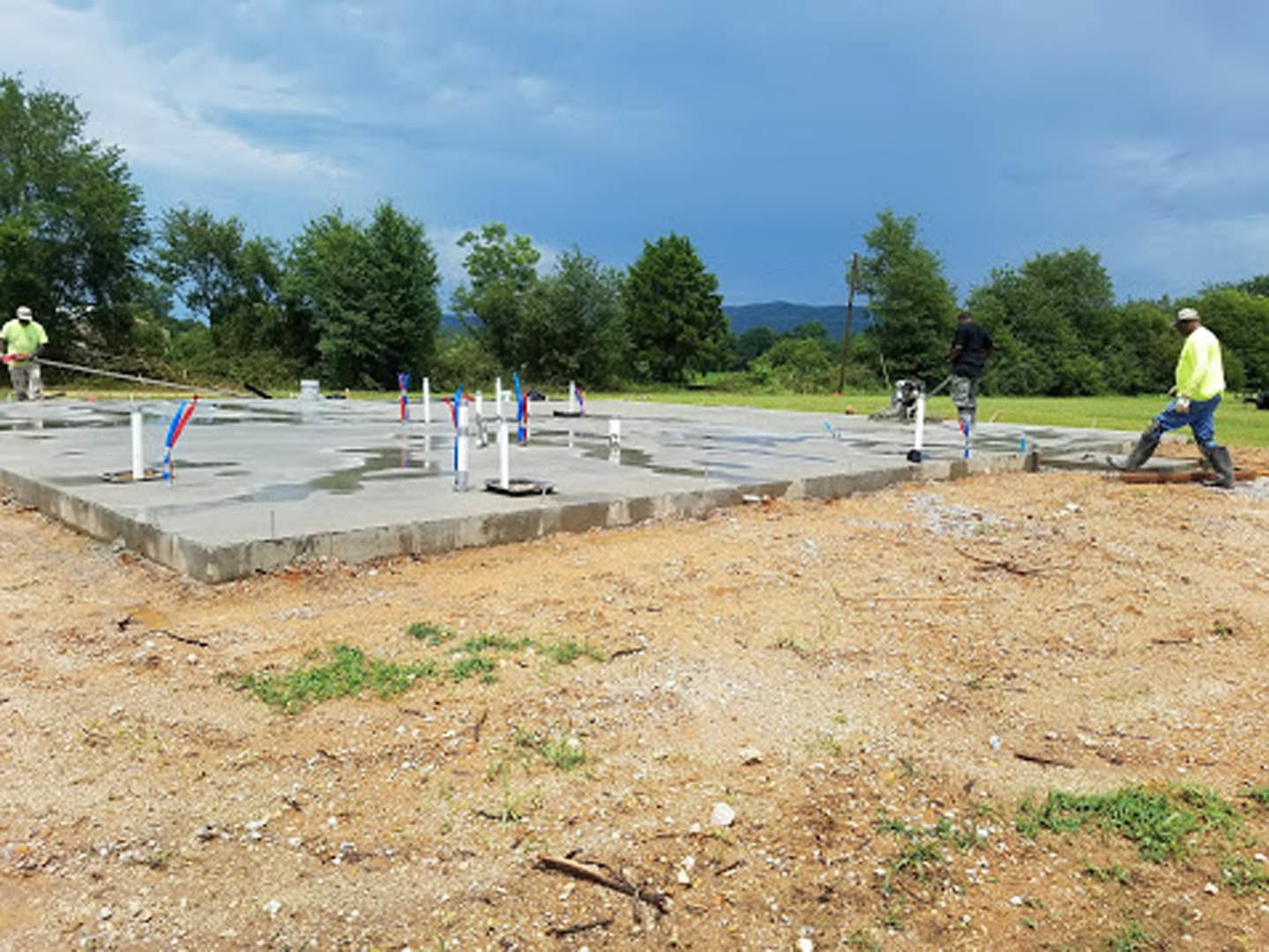 Man in yellow shirt and blue jeans standing on unfinished concrete slab at outdoor construction site, surrounded by grass, soil, and trees under cloudy sky