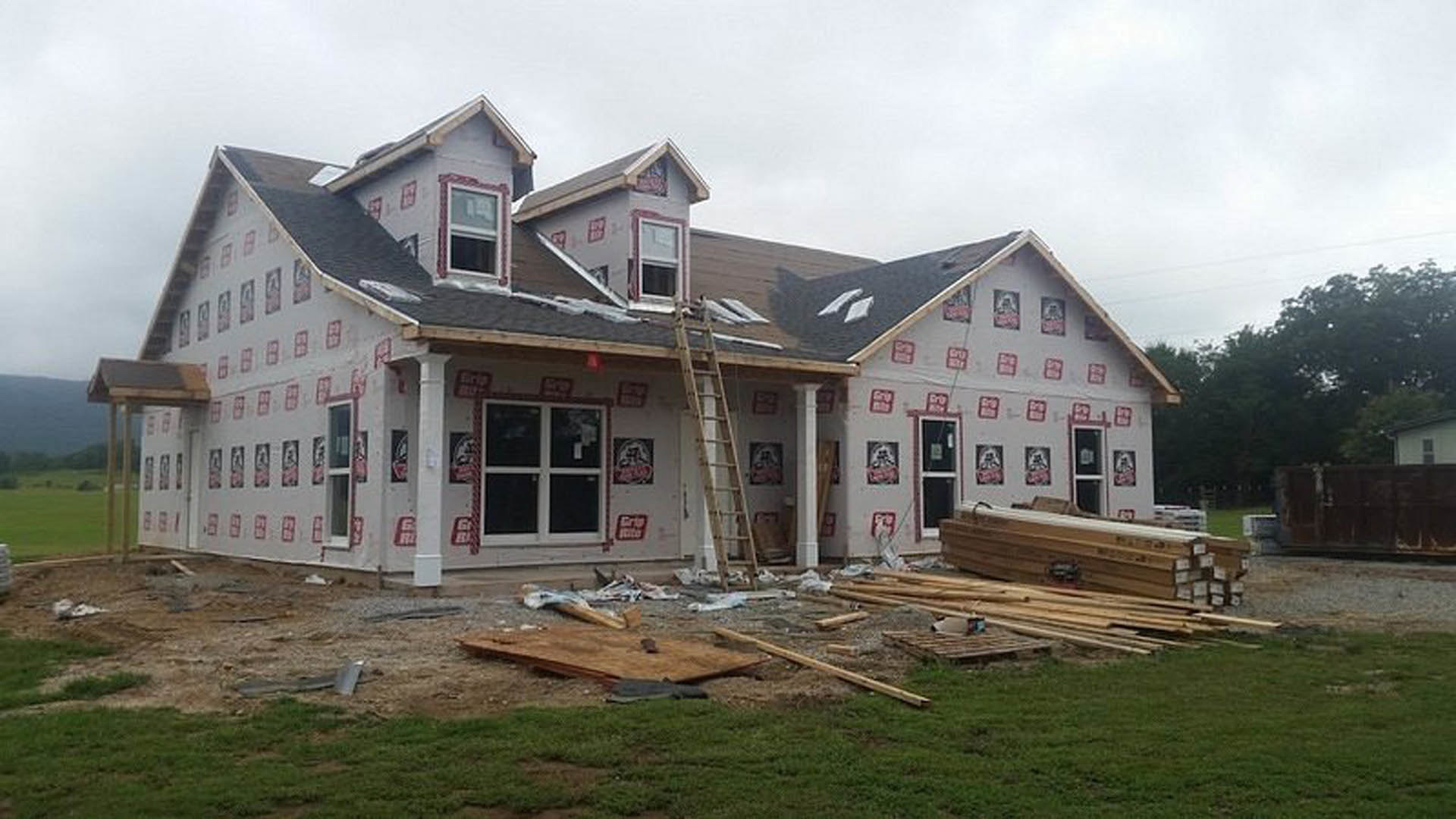 Partially built home with exposed framing, white window frame, ladder leaning against porch, stack of wood planks, light post in front yard, cloudy sky overhead