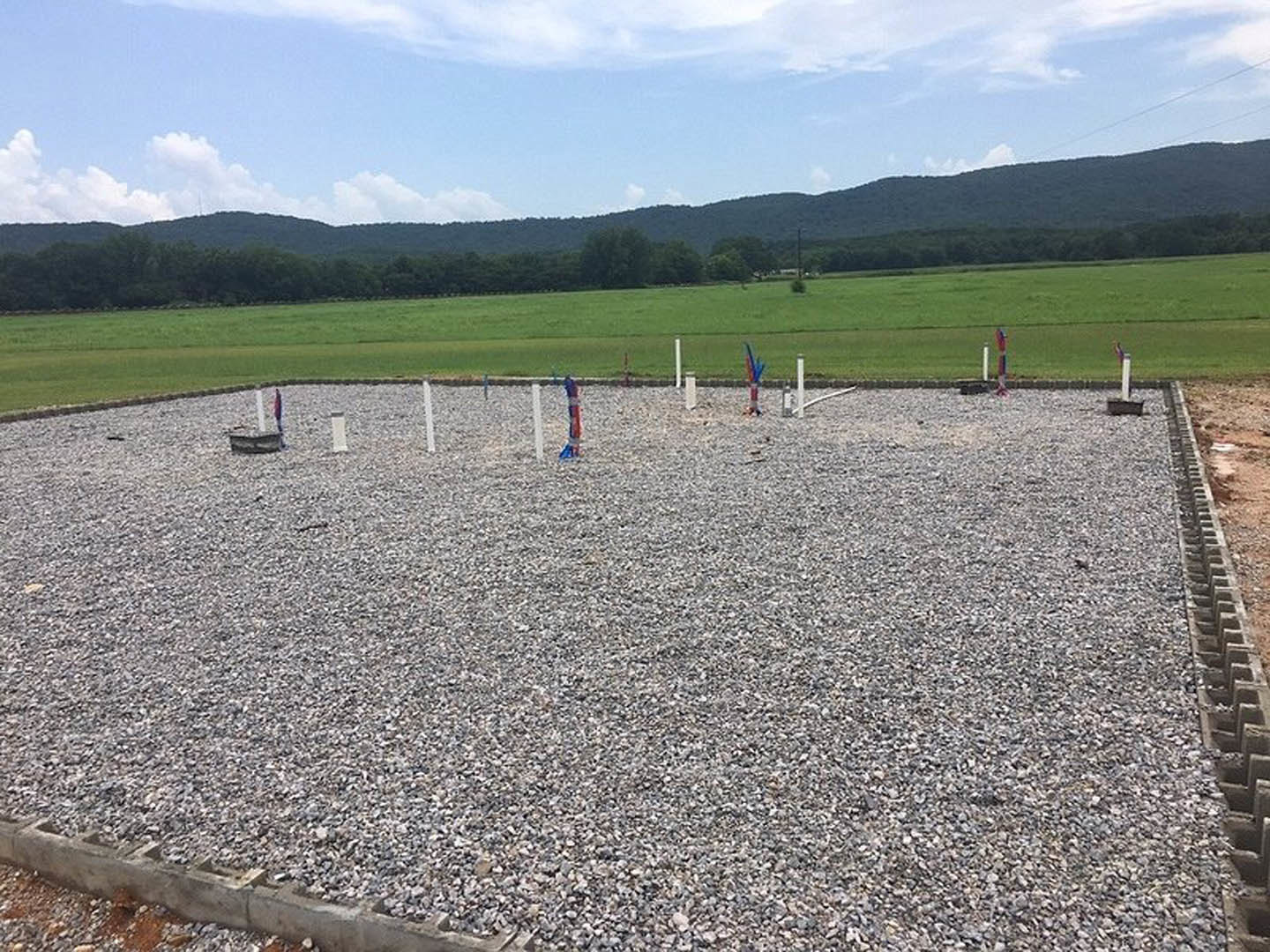 Gravel lot with upright poles scattered across open field, green grass and trees in background, blue sky with clouds overhead, group of people standing near concrete block