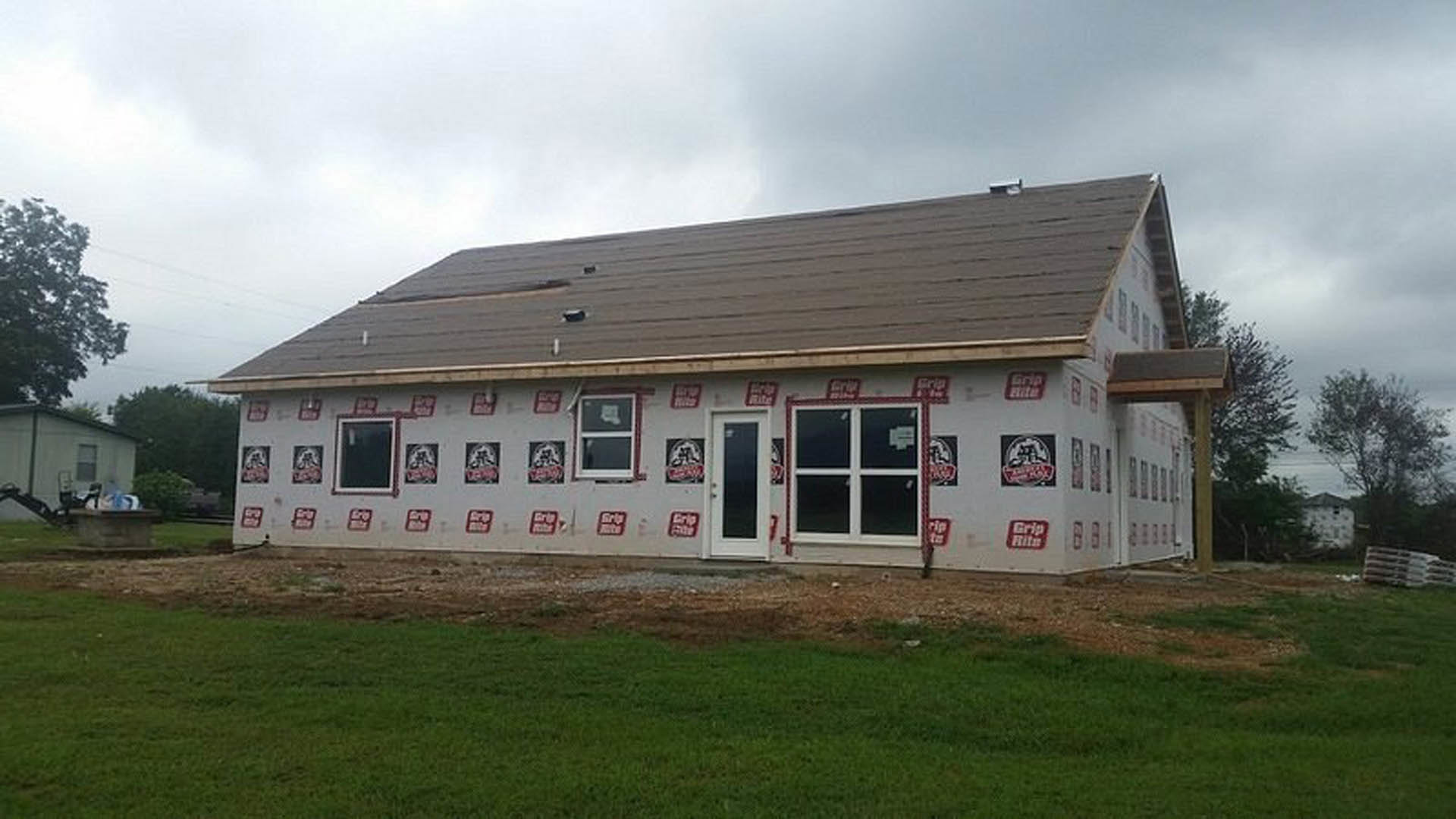 Partially finished house with exposed plywood walls, red and white construction stickers, white-framed windows, door opening, gravel and grass in foreground, cloudy sky above
