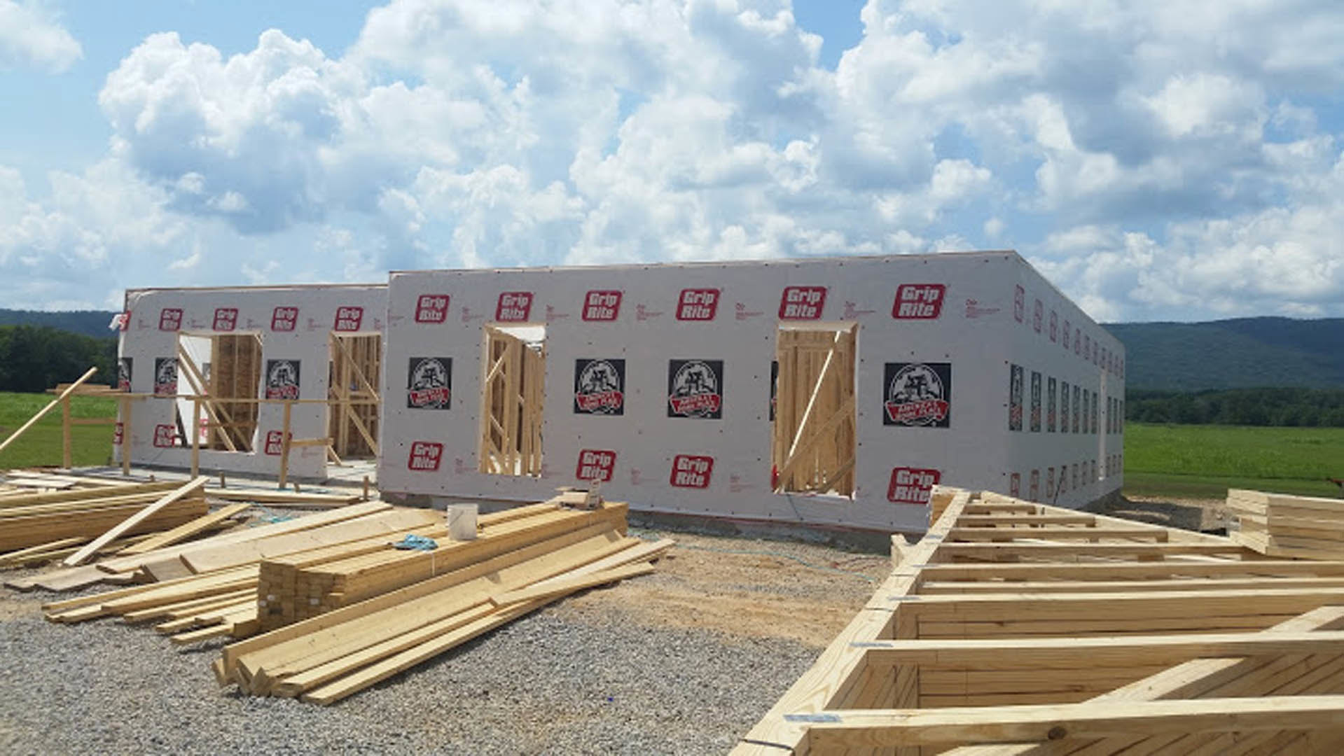 Wood-framed exterior walls and beams under construction, exposed lumber, scattered wood planks on ground, partially built wooden structure, blue sky with clouds overhead