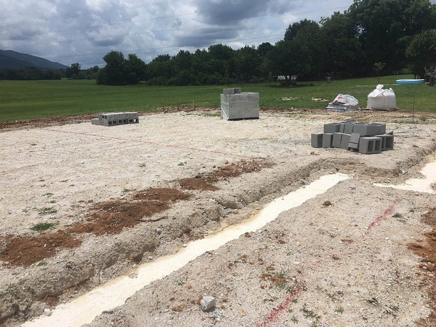 Grassy construction site bordered by trees, scattered grey concrete blocks, dirt ditch, and white bag on lawn