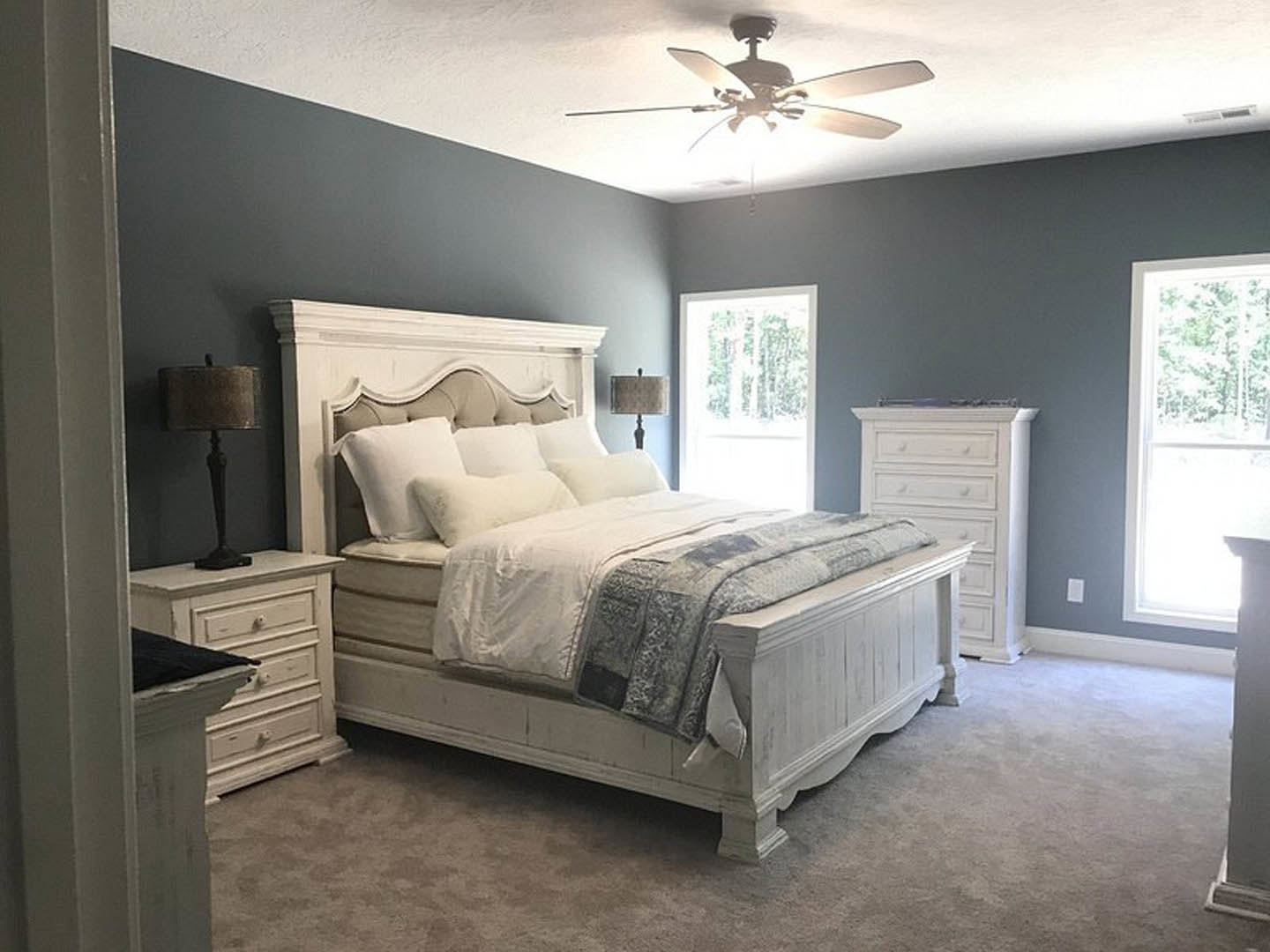 Bedroom with white bedding, ceiling fan with light, white dresser beside bed, lamp with shade on nightstand, window with white frame, neutral walls and flooring