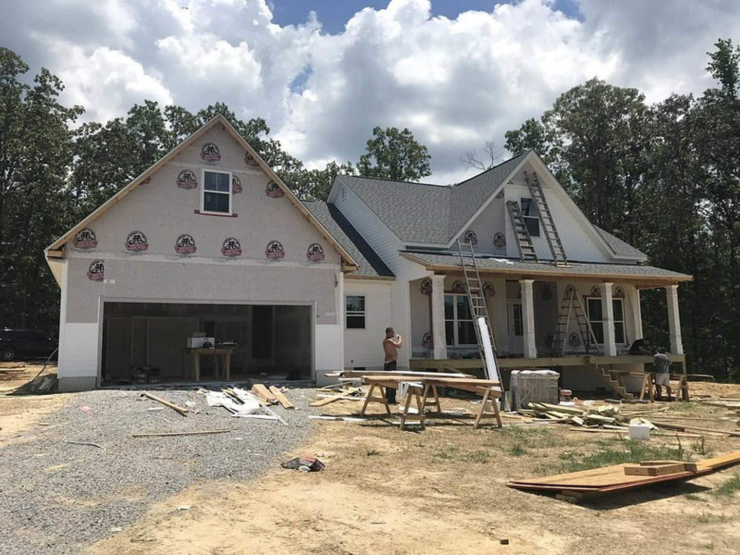 Partially built house with exposed wooden framing, white-framed windows, attached garage, man painting exterior wall, cloudy sky overhead, construction materials scattered on