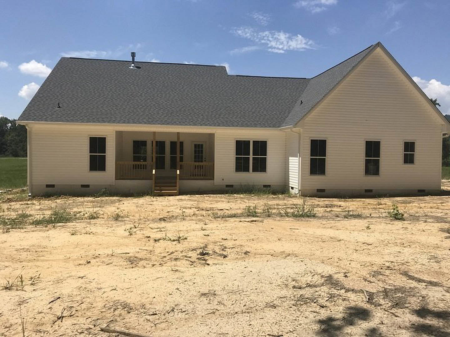 Two-story house with beige siding, white trim windows, covered front porch, and dirt yard with sparse grass under a cloudy sky