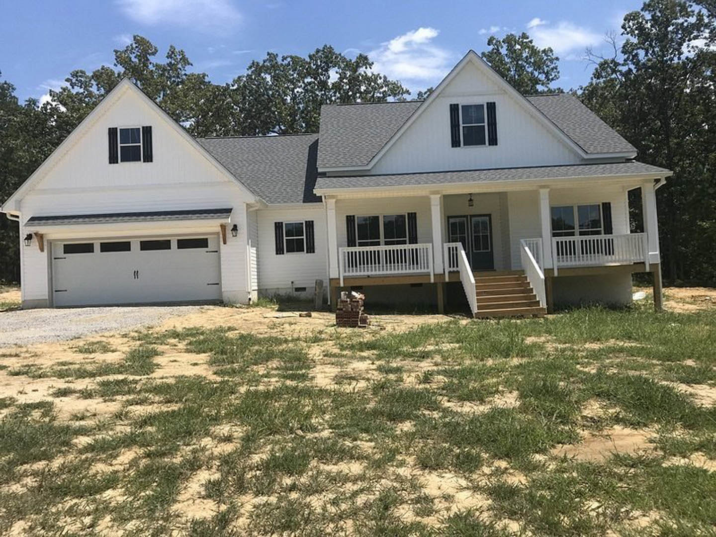 White two-story house with covered porch, paved driveway, green lawn, and stacked bricks near the entrance; large windows with white frames and mature trees in the yard.