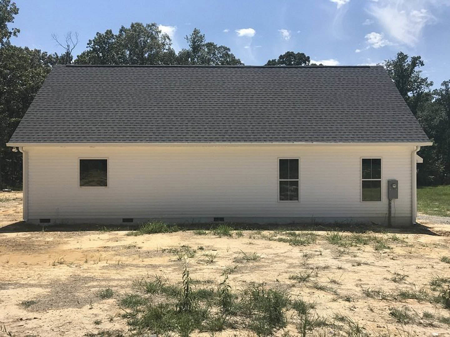 White cottage-style house with gabled roof, large window framed in white, wooden fence in front, grassy yard with patches of dirt, trees and cloudy sky in background