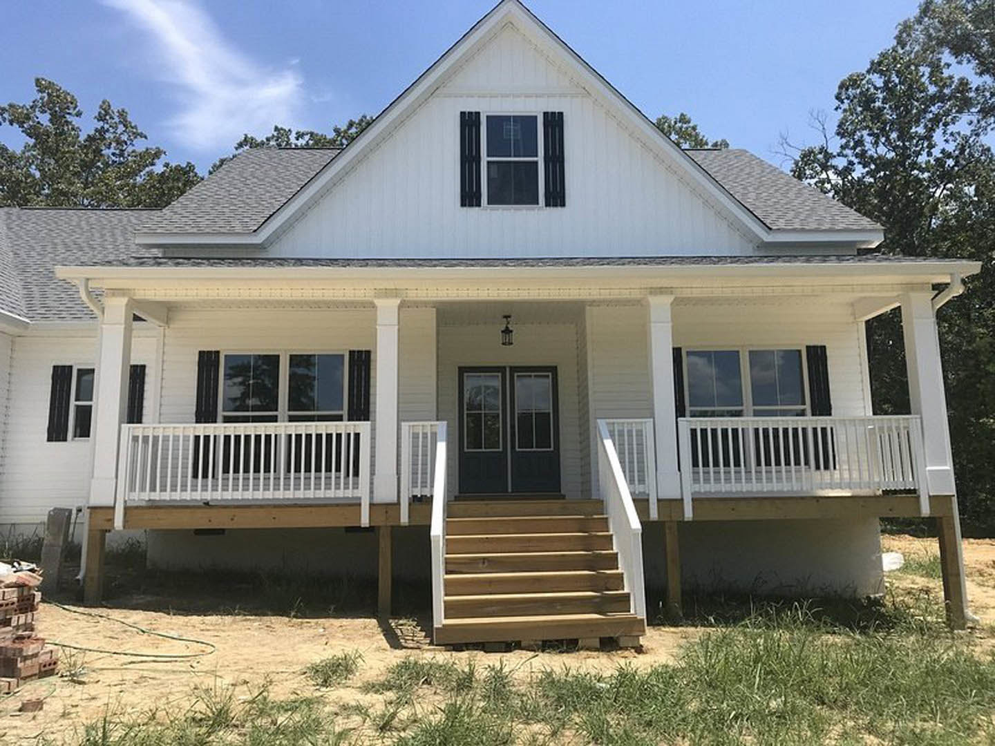 White siding house with covered porch, wooden stairs, double glass doors, and large windows; green grass surrounds sandy walkway.