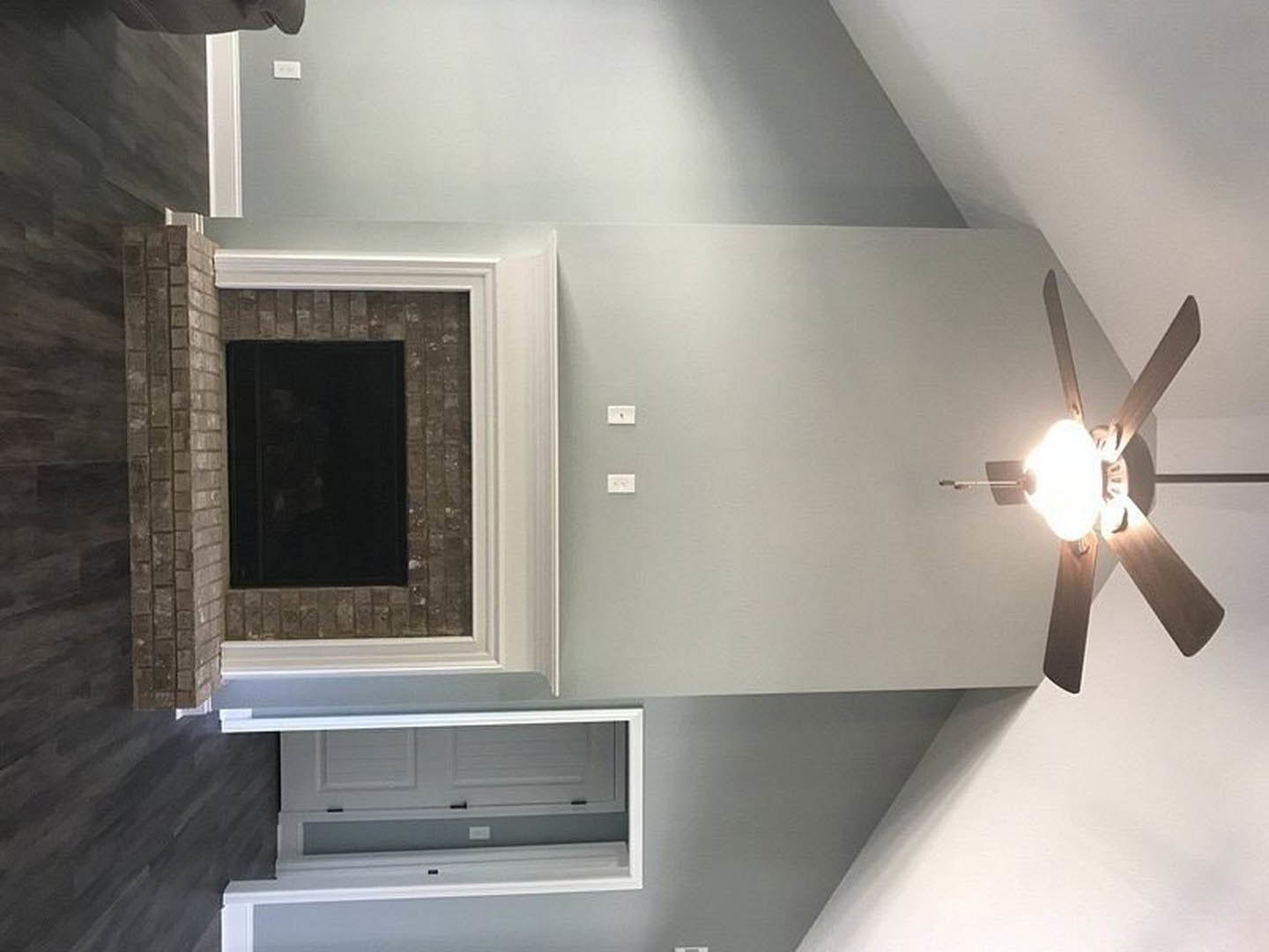 Ceiling fan with light fixture above living room featuring brick fireplace, white cabinetry, plaster walls, and wood flooring