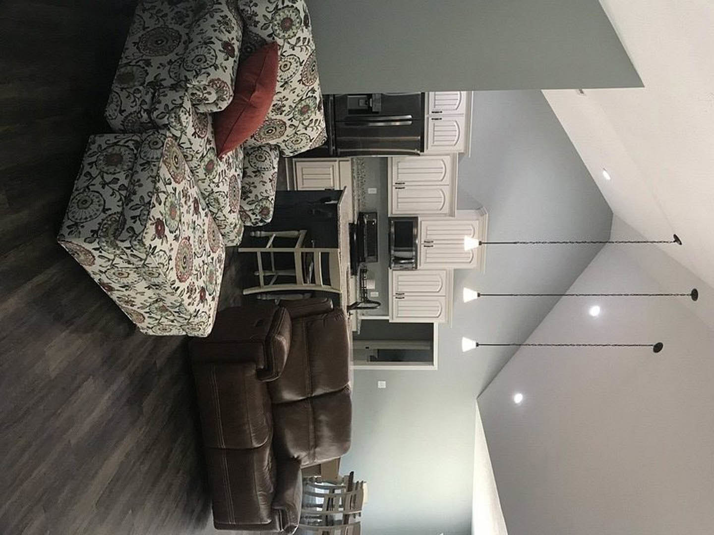 Brown leather couch with white and red pillows in open living room, adjacent to modern kitchen; ceiling lights, two lamps, and white shelving visible.