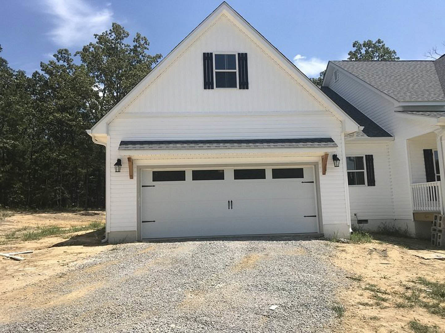 White house with attached garage, white-framed windows, gravel driveway, and trees in the background
