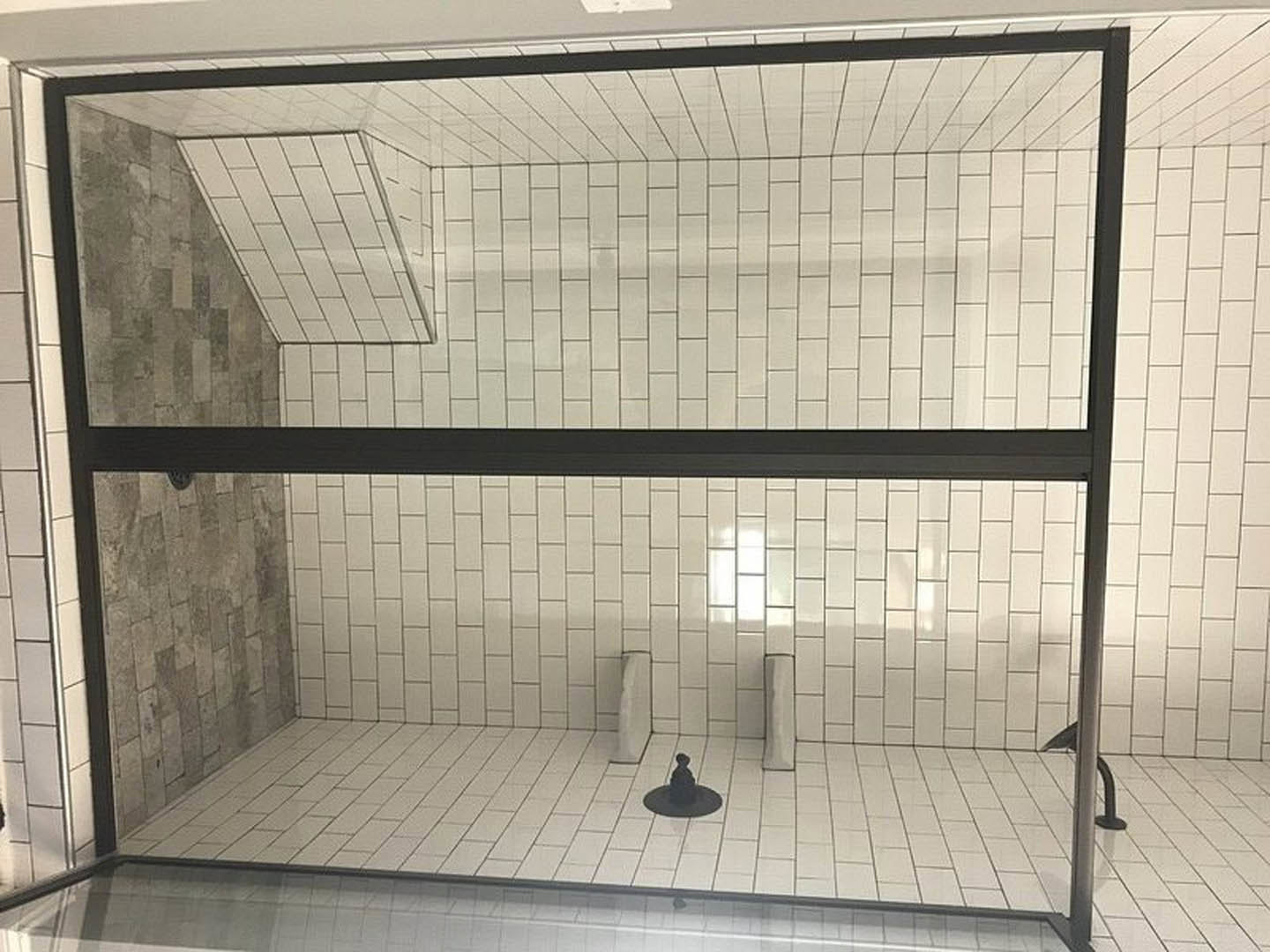 Glass shower door with chrome handle, white tile walls and floor, black border accent, black hat resting on tile surface