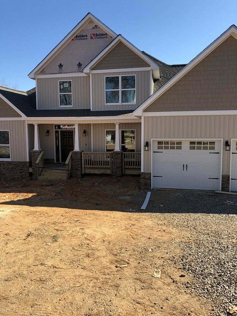 Two-story home with gray siding, attached garage with white paneled door, concrete driveway, covered porch featuring wooden railing, white-trimmed windows, and manicured lawn.