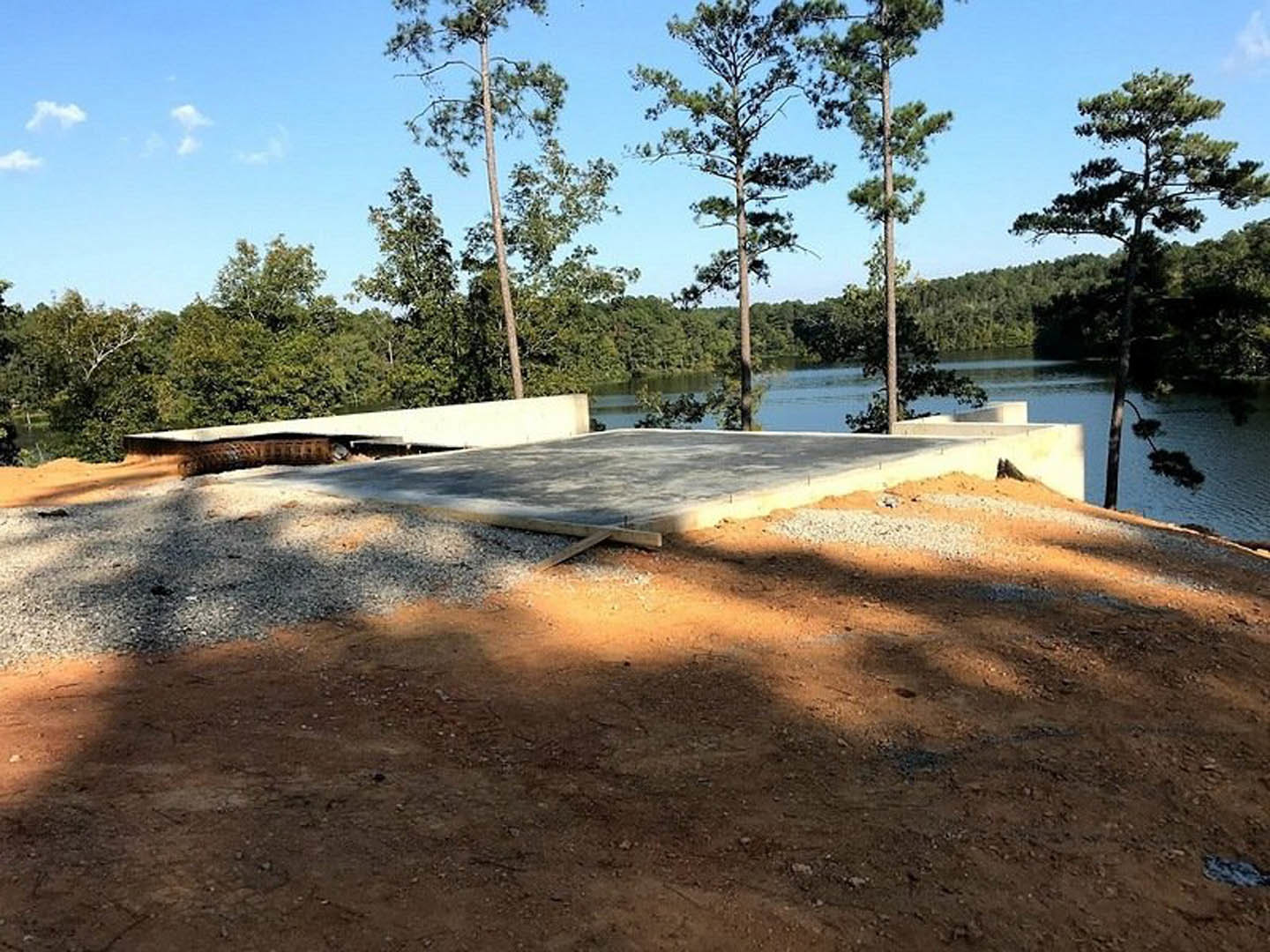 Concrete slab with a snake resting beside a lake, surrounded by tall trees and a dirt surface, under a partly cloudy sky