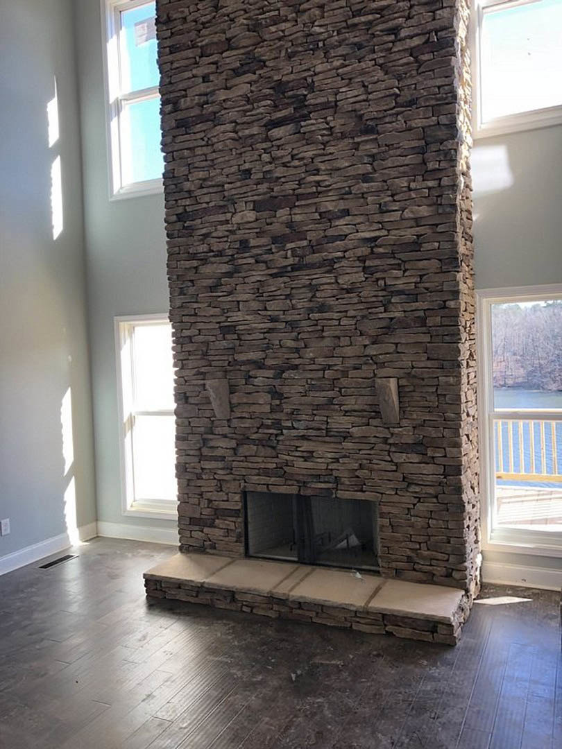 Stone fireplace with glass door set in a brick accent wall, adjacent to a window and white door, dark flooring and stone step visible in spacious indoor room.