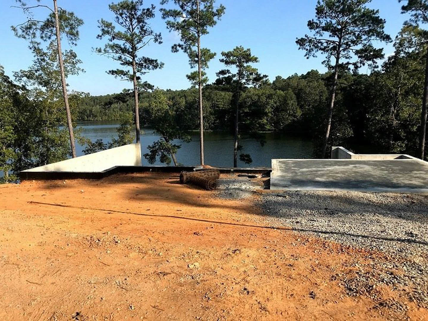 Partially built custom home with exposed wood framing, dirt ground scattered with construction materials, tall leafy trees surrounding the site, and a white roof visible against a