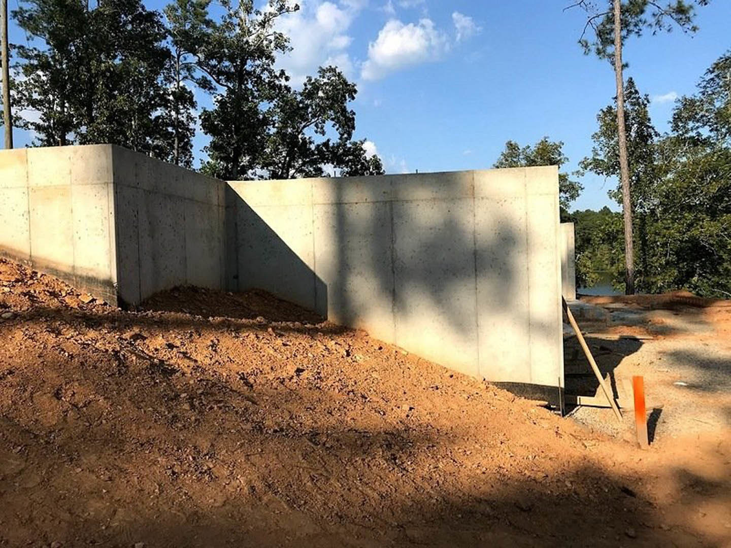 Concrete retaining wall bordered by dirt, tall tree trunks, and blue sky with scattered clouds; orange pole and wooden stick embedded in ground