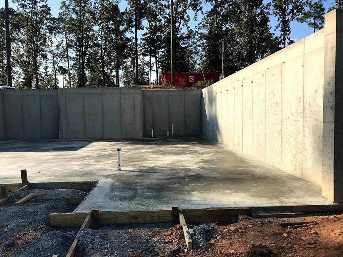 Concrete foundation with vertical support poles, surrounded by tall trees in a wooded area, close-up of unfinished concrete wall with holes, red fence featuring a white sign, wall