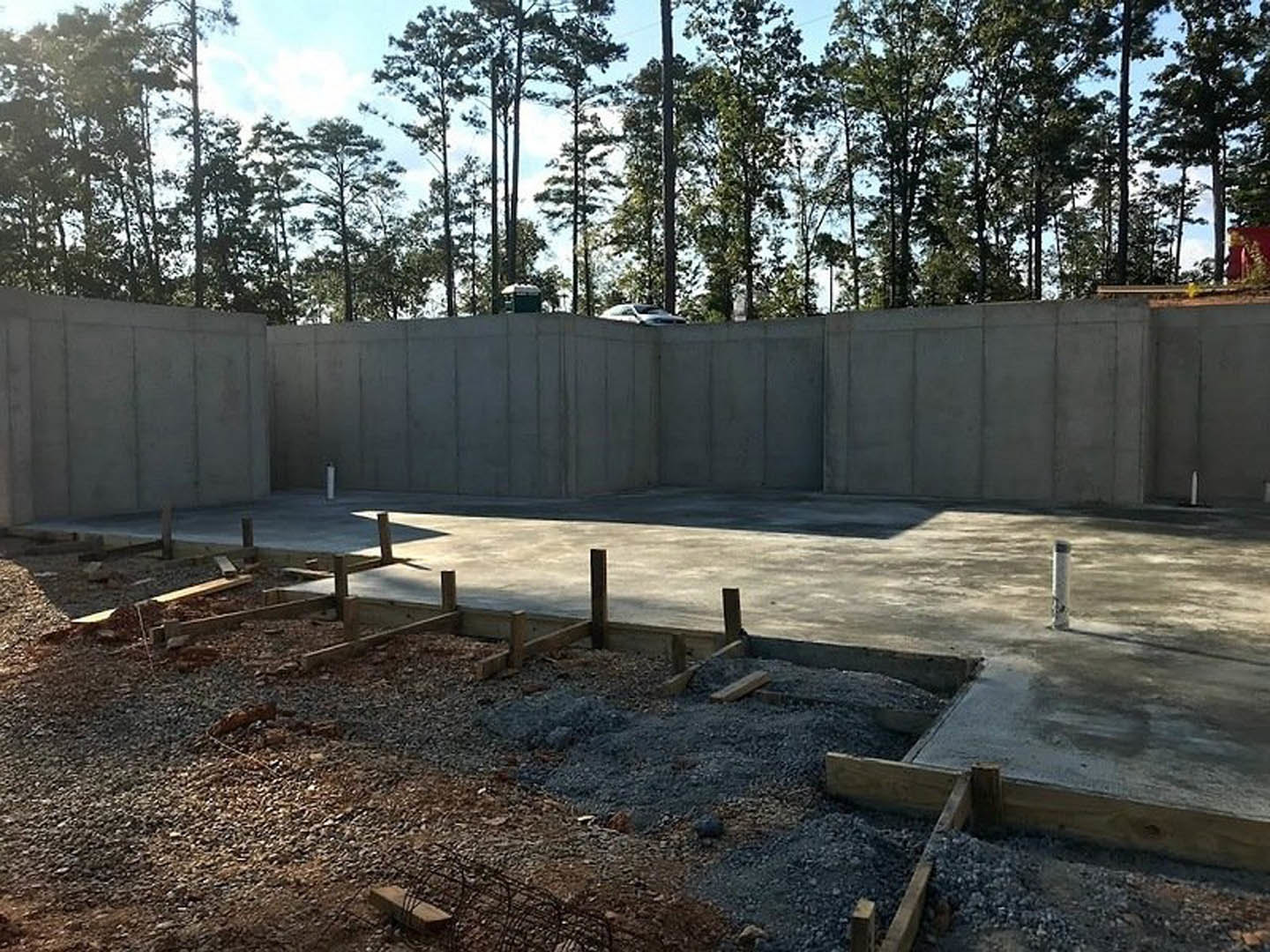 Concrete foundation with wood framing, gravel construction site, parked car atop concrete wall, tall trees in background, blurry figures near wooden posts