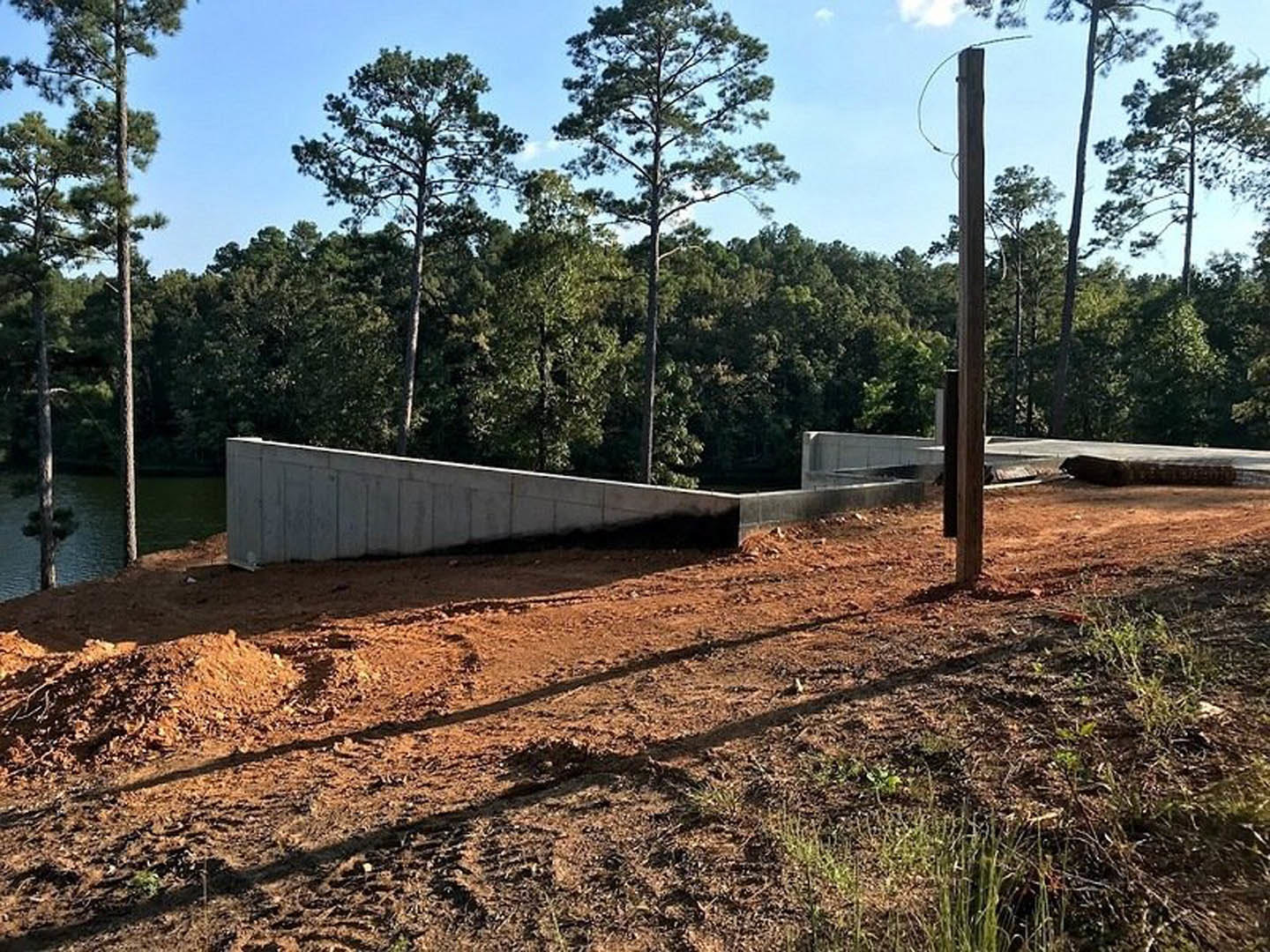 Dirt and grass construction site bordered by a long grey basement wall, tall trees with many branches in the background, close-up of a pole in foreground