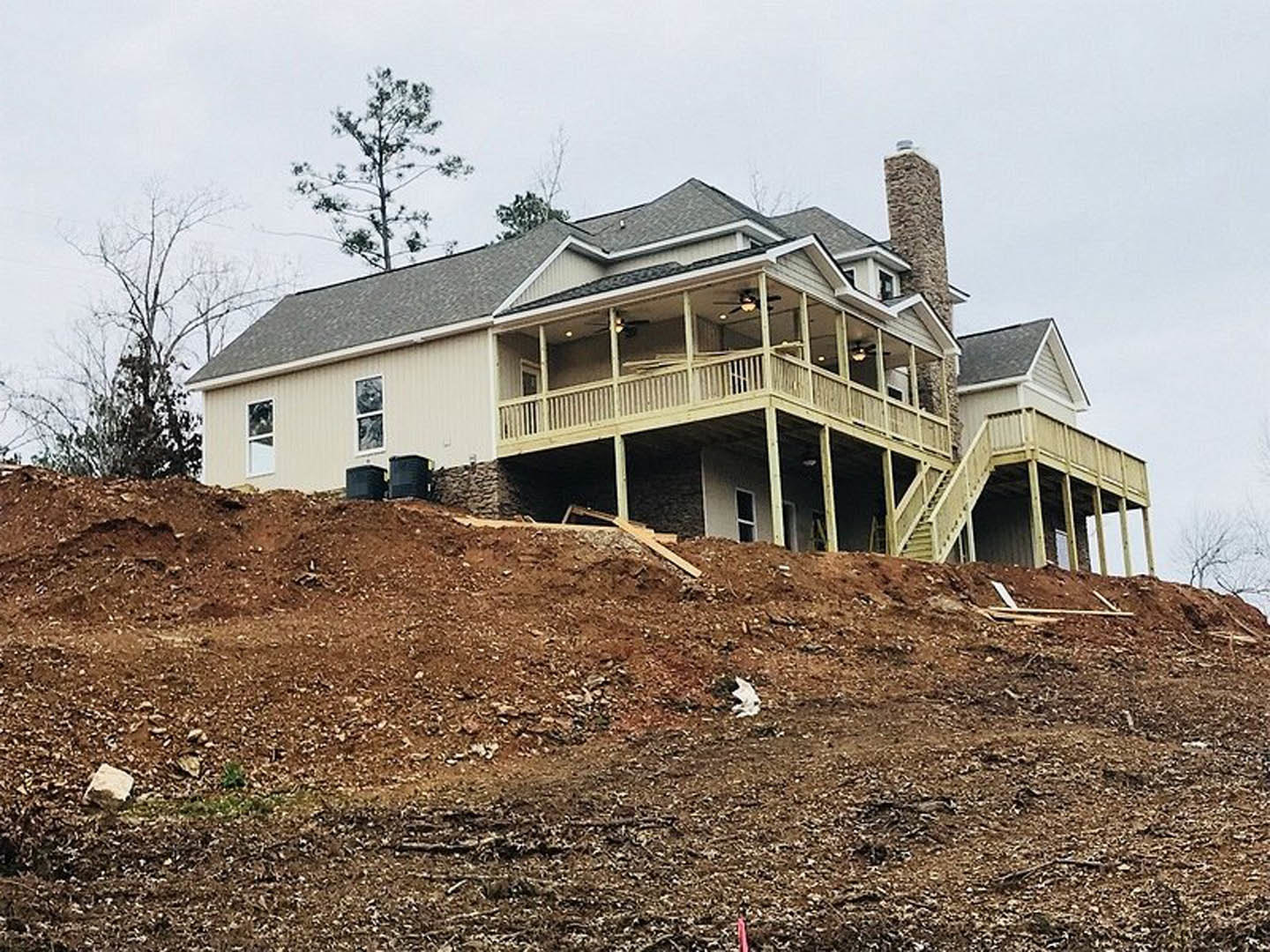 Modern two-story home with stone chimney, large windows, and wooden balcony, situated on a grassy hill surrounded by trees under a clear sky