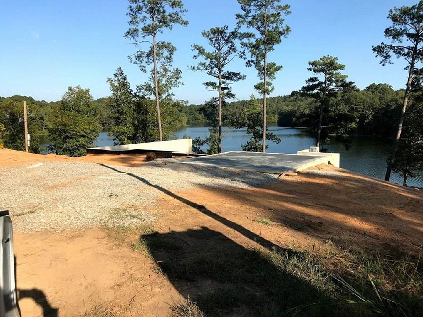 Modern lakeside home with light stone exterior, expansive windows, and shaded lawn; mature trees frame the property, a wooden bridge crosses the water, and reflections ripple