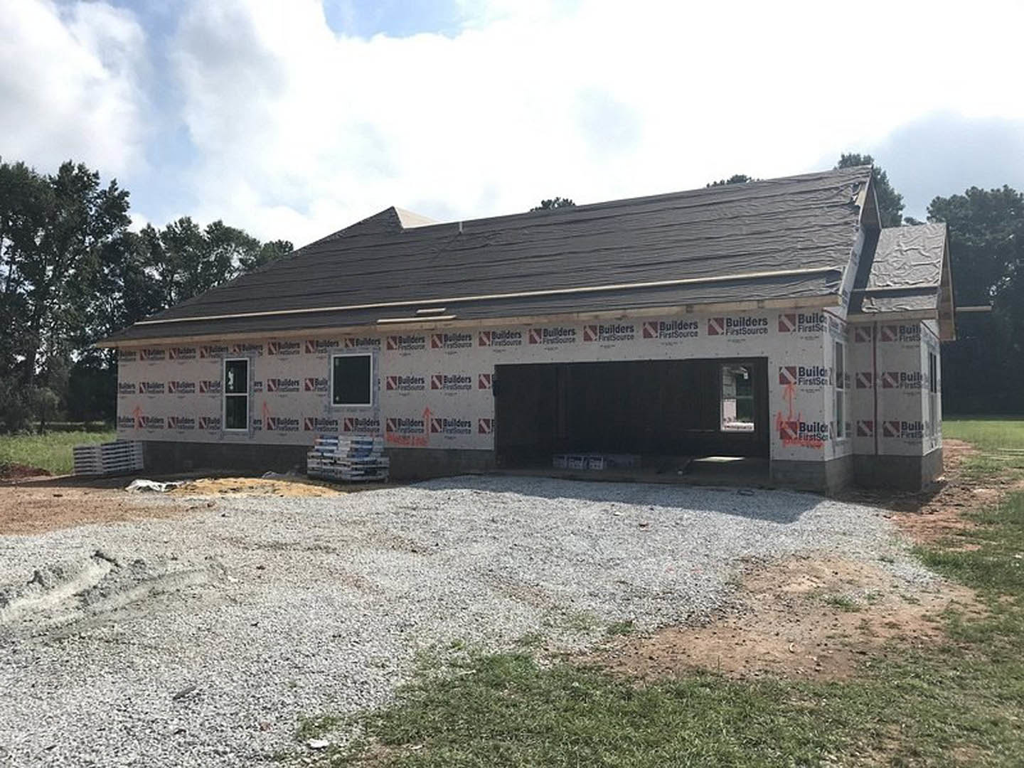 Modern house under construction with gravel covering the front yard, attached garage featuring a black door with white trim, scattered pallets on the lot, and trees in the