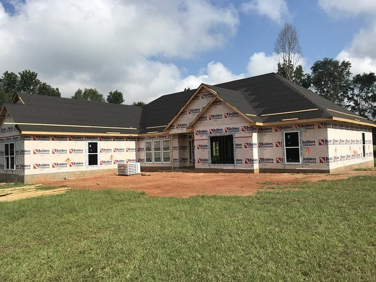 Partially built house with black roof, white-framed windows and door, surrounded by dirt patches and green lawn under cloudy sky