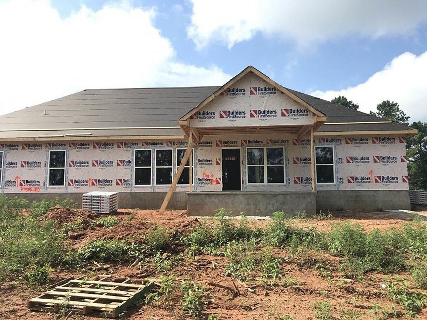 Modern home under construction with exposed framing, patch of dirt in foreground, wooden pallets stacked near porch, ladder leaning against exterior, white-framed window, black