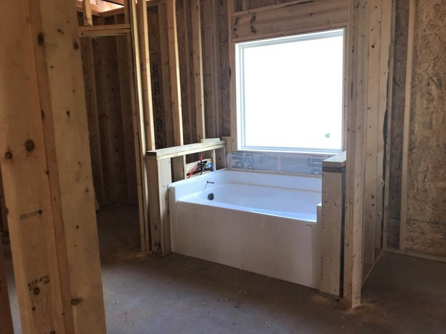 Freestanding white bathtub in unfinished bathroom with exposed plaster walls, wooden-framed window, and visible construction details