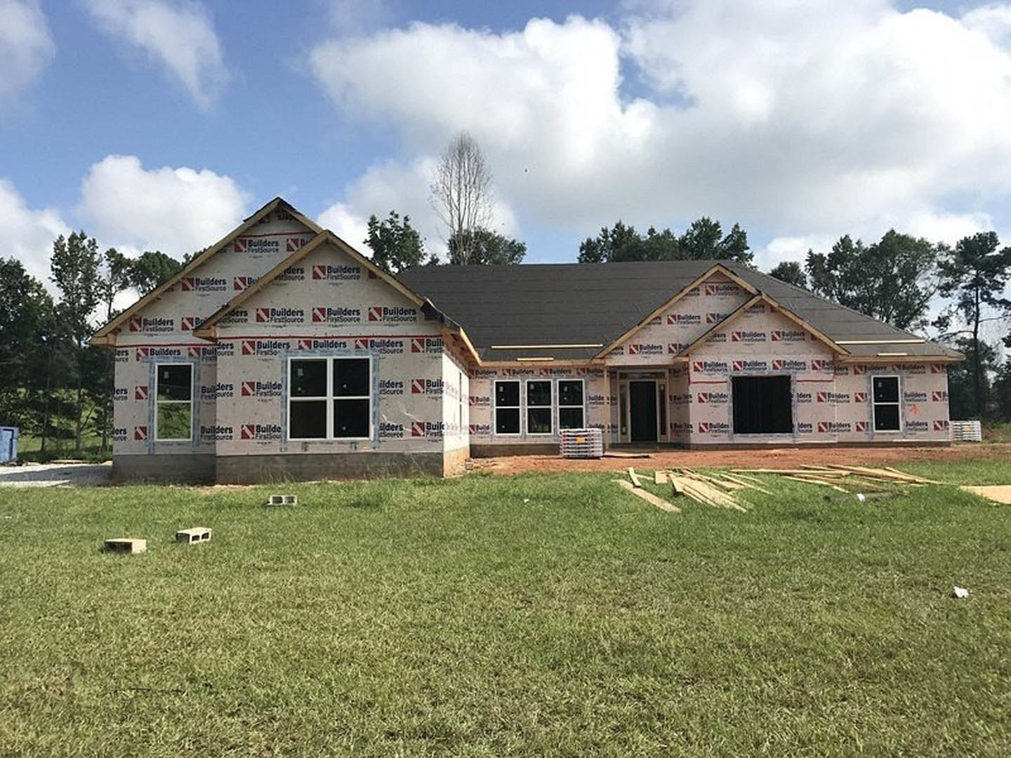 Two-story farmhouse under construction with unfinished exterior walls, large windows, and a green lawn in front; construction materials and pallets scattered near the foundation.