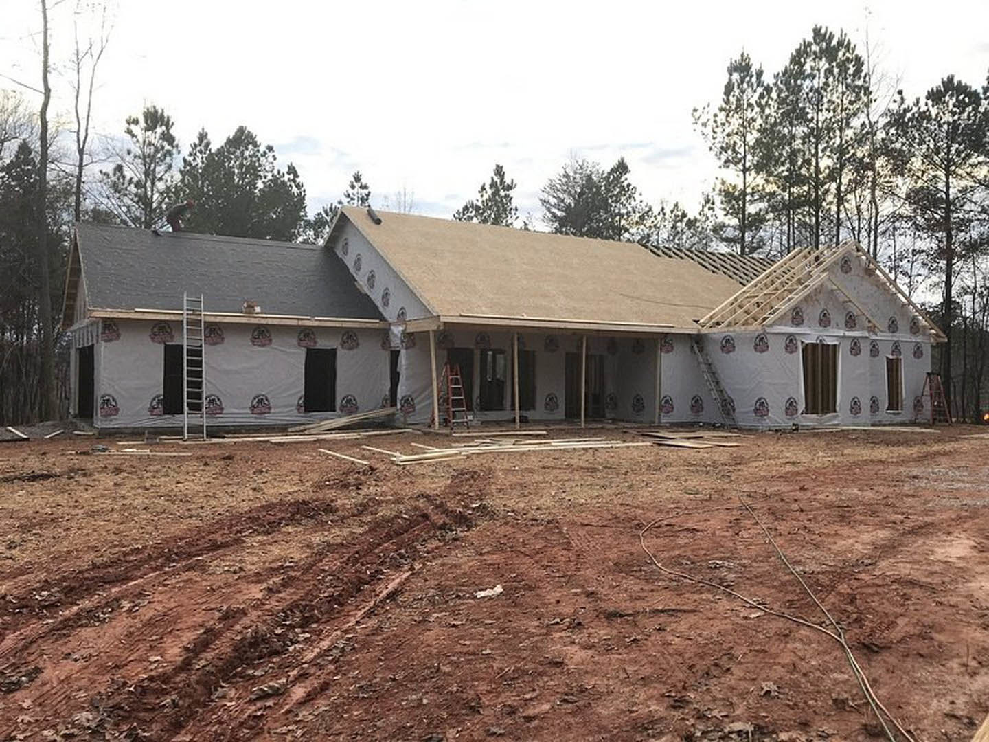 Framed house under construction with exposed wood beams, white ladder leaning against unfinished wall, dirt ground scattered with lumber, partially visible roof, trees in