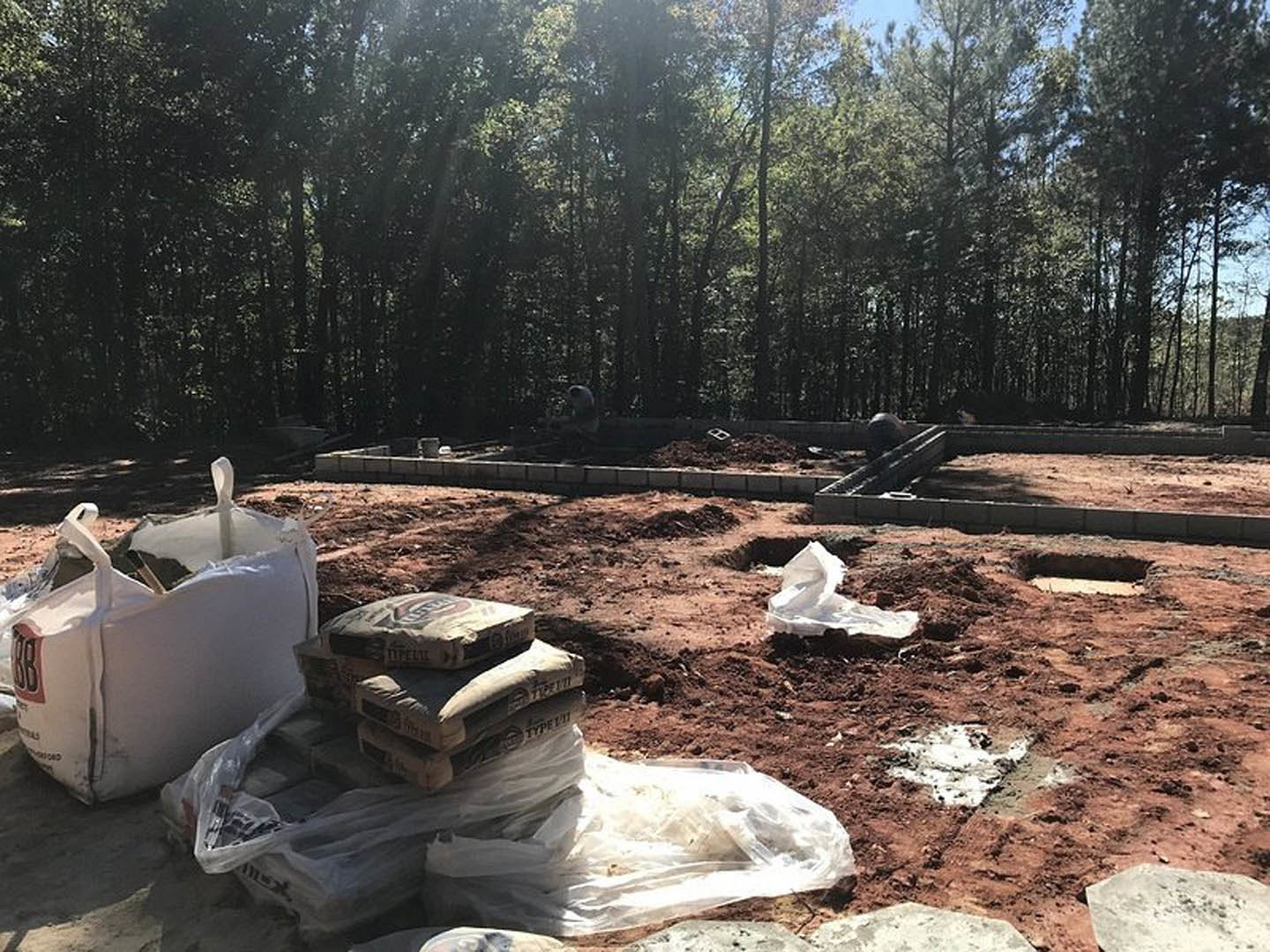Construction site with stacked cement bags and bricks on bare soil, surrounded by trees and scattered rocks