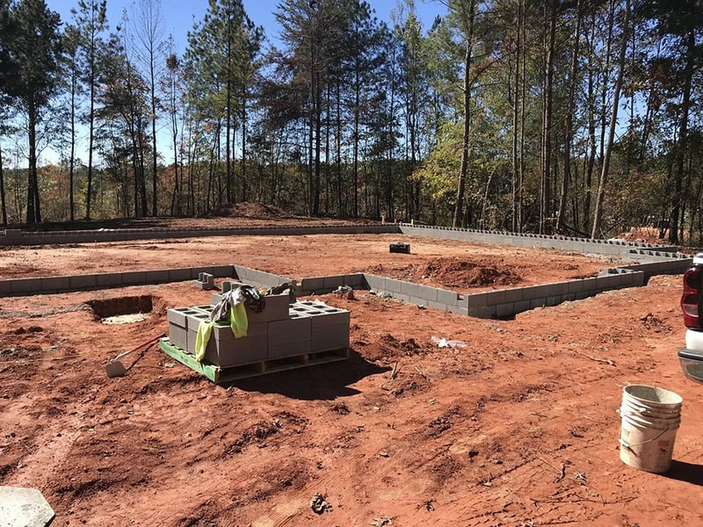 Stacked grey bricks on a construction site with exposed soil, scattered pallets, and surrounding trees in the background