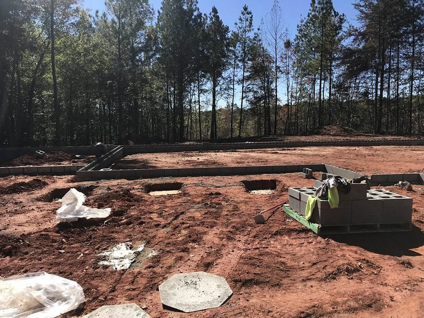 Grey brick pallets and scattered construction materials on bare soil, white square marking foundation area, forested tree line in background