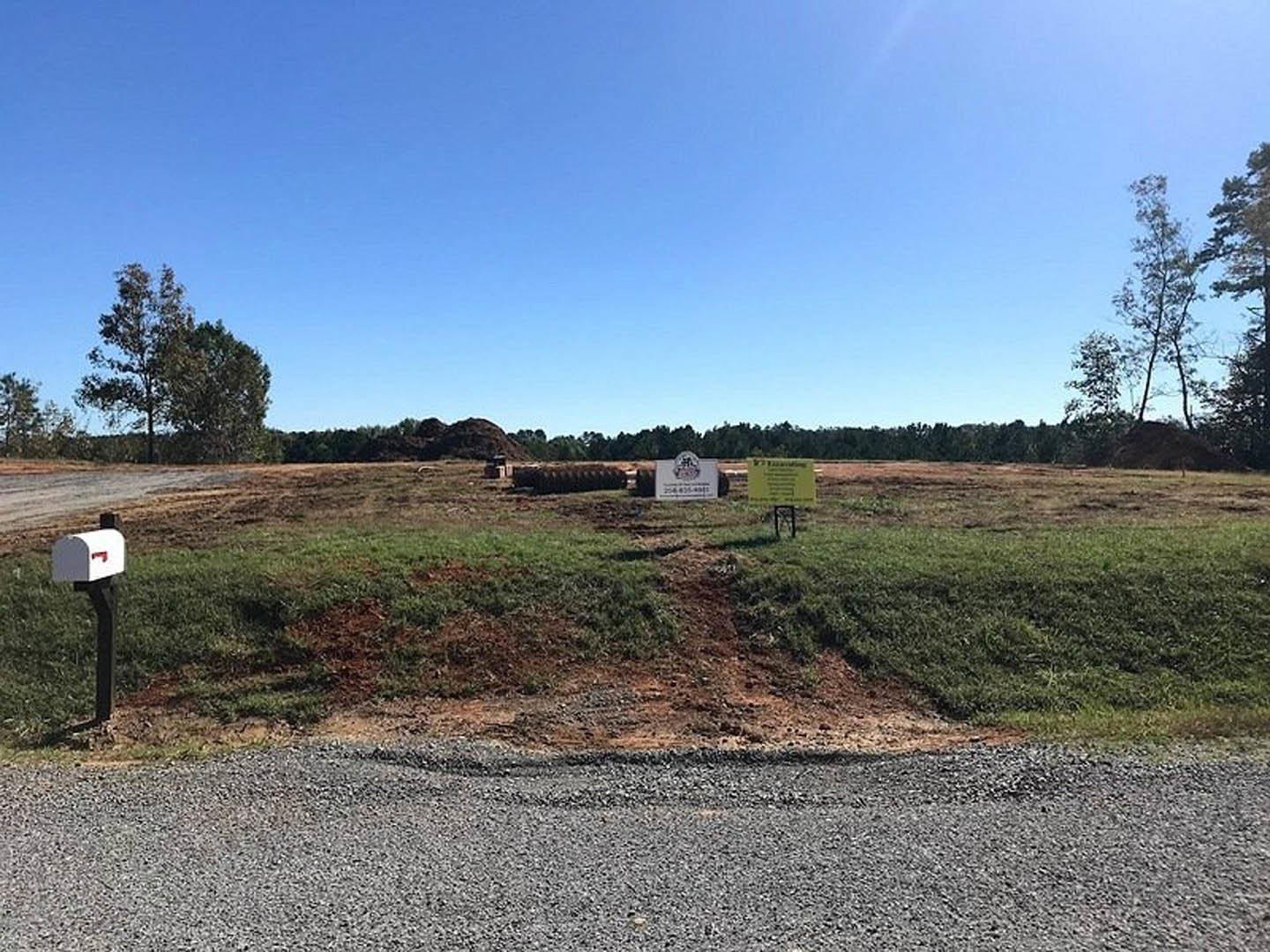 White rectangular sign mounted in grassy field with gravel road, leafy tree, and blue sky with scattered clouds in background.