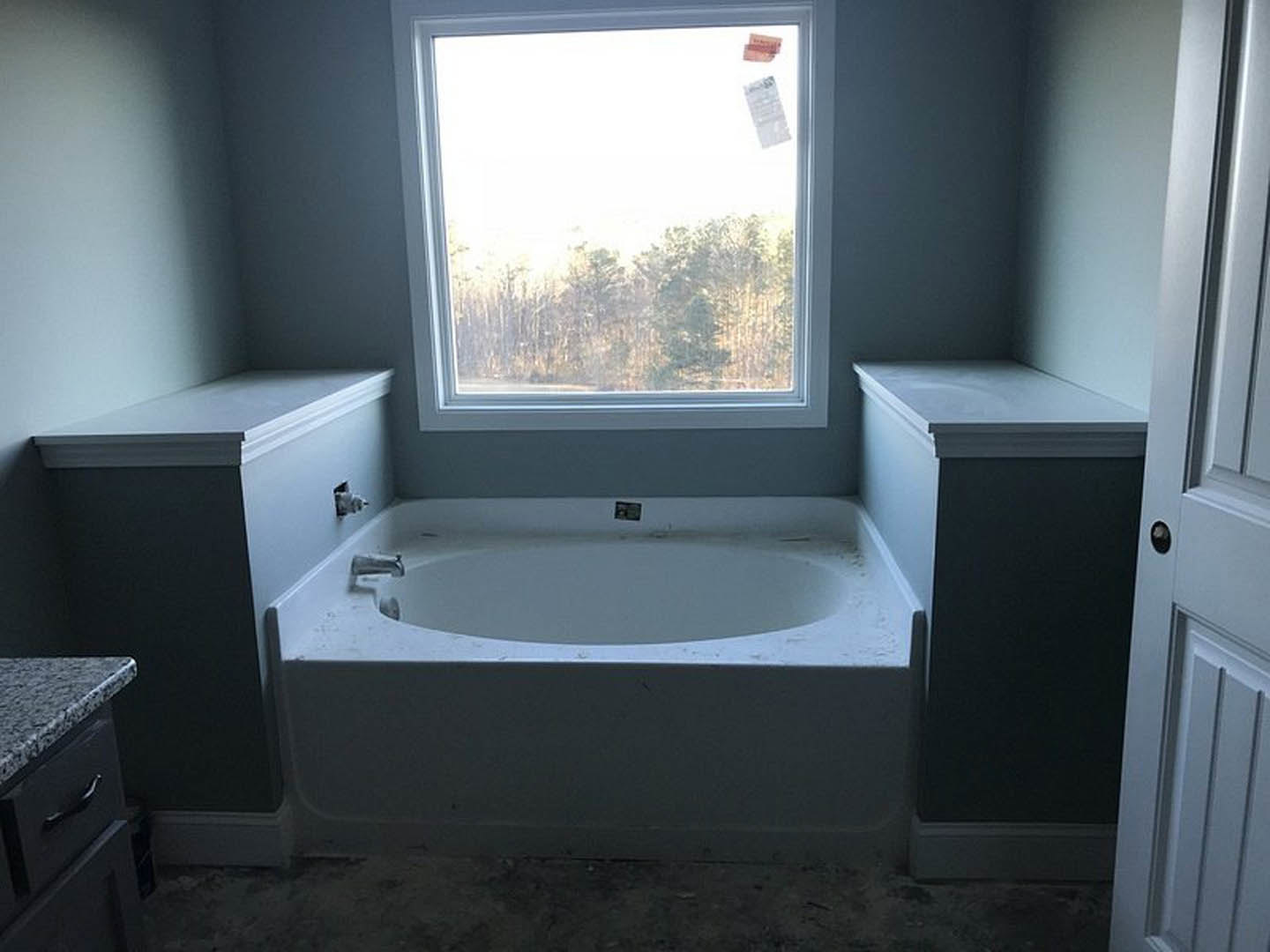 Freestanding white bathtub with chrome faucet, gray tile walls, large window, wall-mounted mirror, and modern bathroom accessories